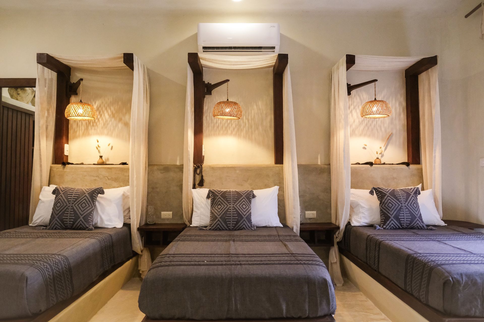 Three beds with canopies, gray bedding, woven lamps, neutral-toned walls, air conditioner.