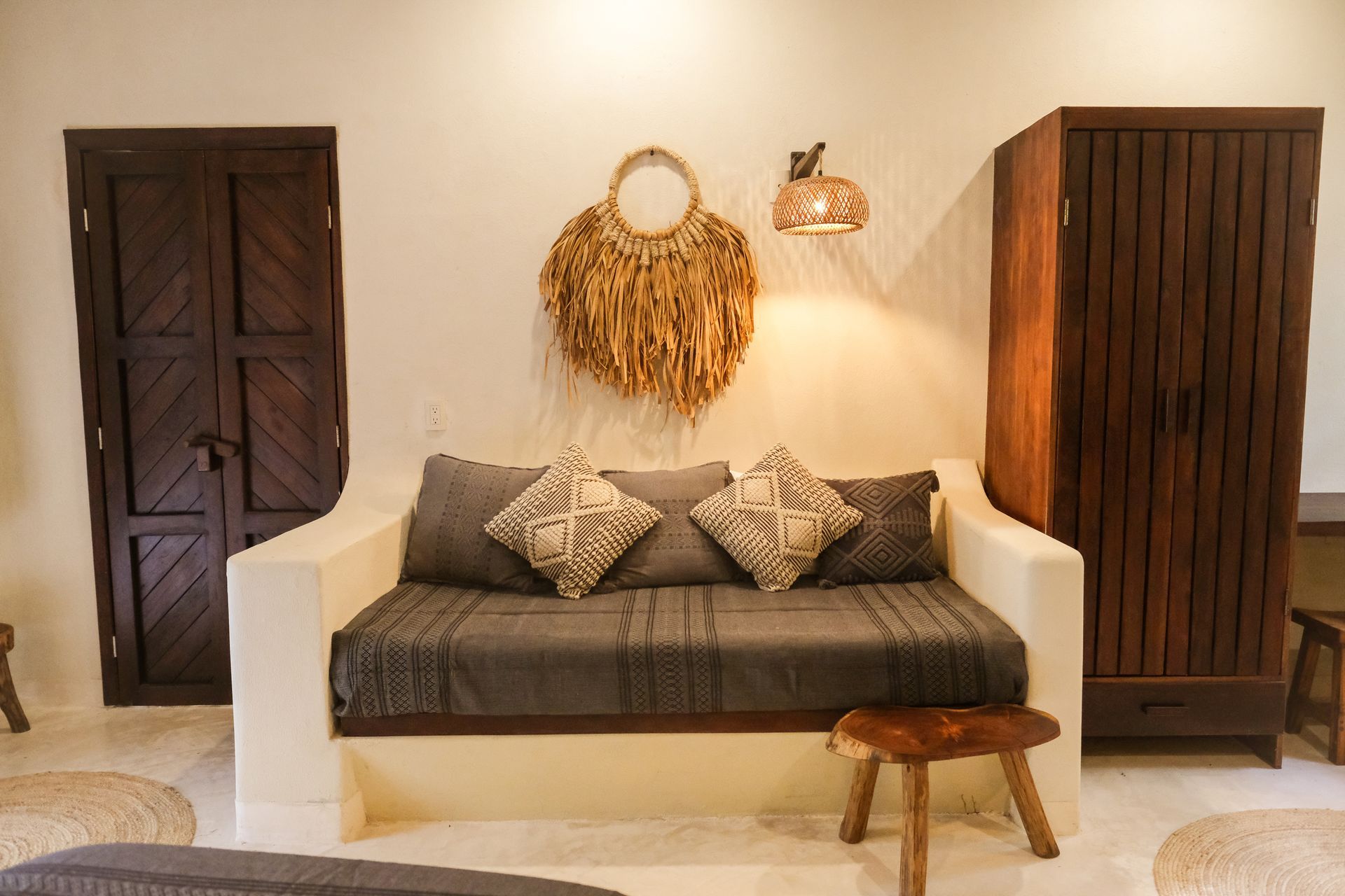 White-walled room with dark wood furniture: sofa, armoire, and door. Decorative woven items, gray cushions.