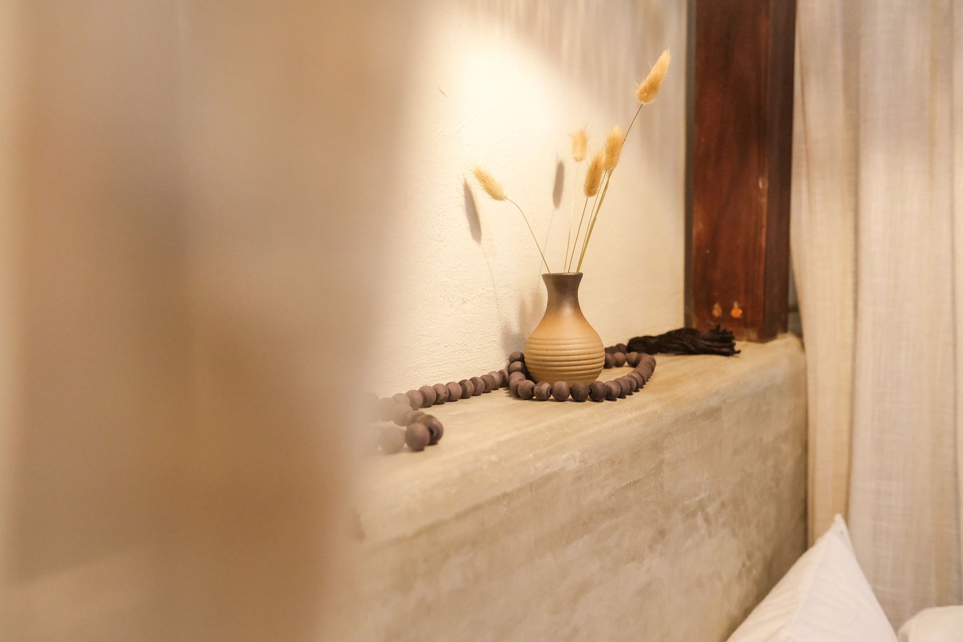 A vase with dried flowers sits on a shelf with beads, next to a wood pillar and curtains.