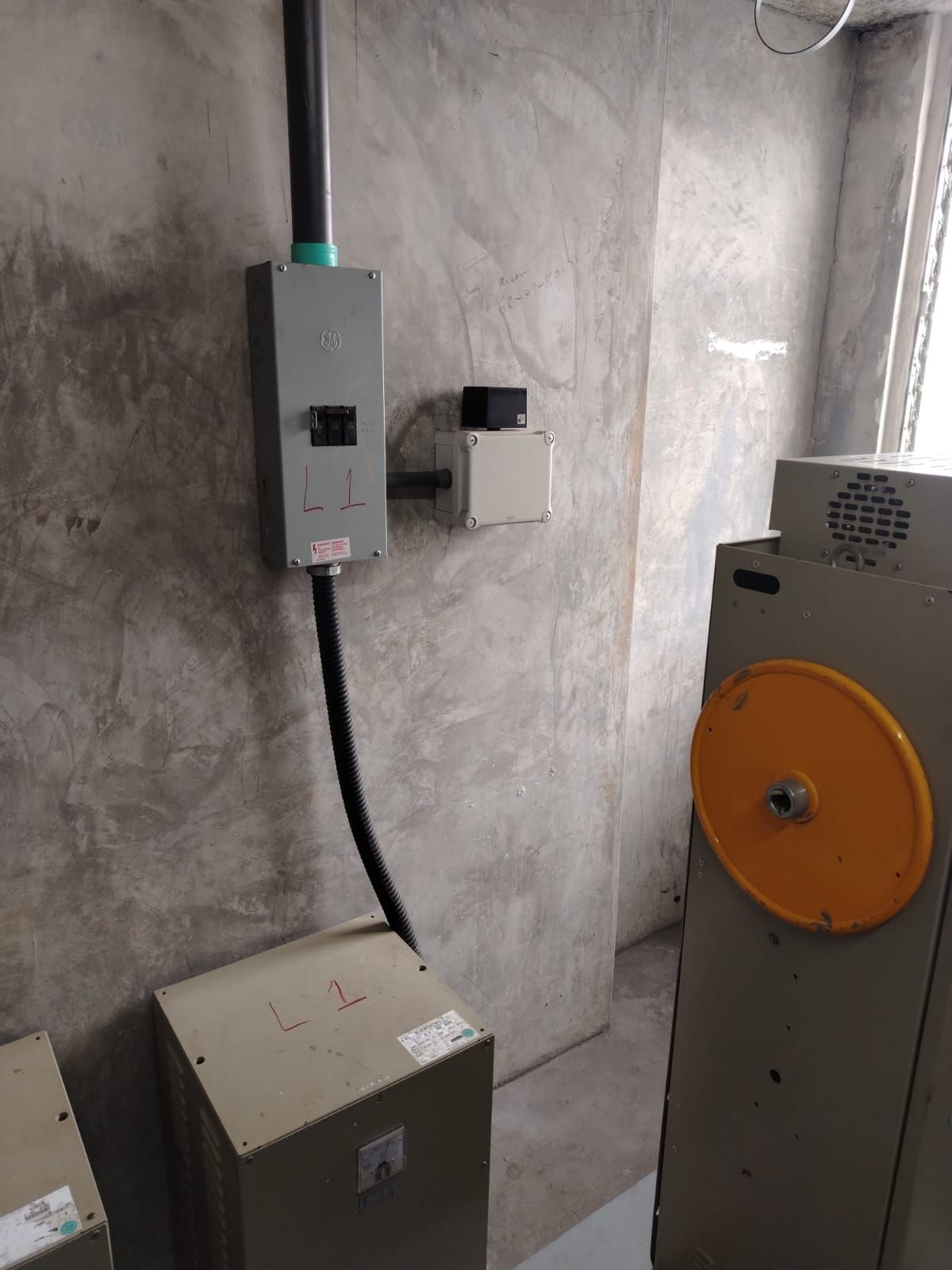 Caja eléctrica y conducto en una pared de hormigón texturizado, junto a maquinaria y otra caja.