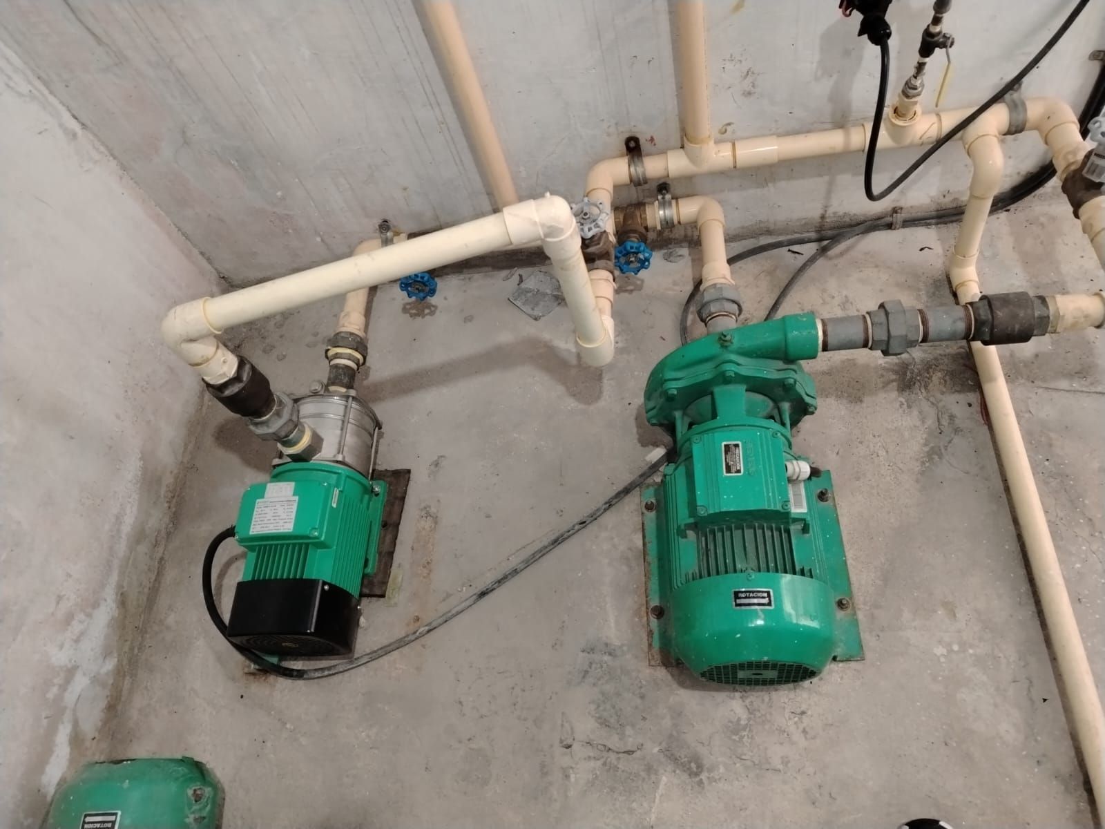 Dos bombas de agua verdes con tuberías color beige adosadas a una pared de hormigón.