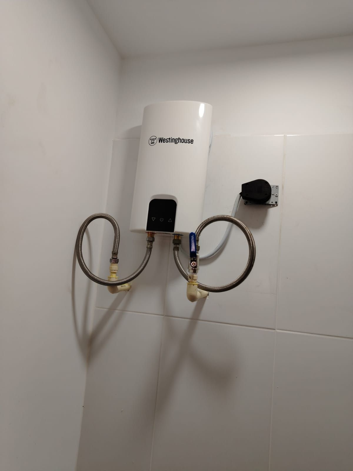 Calentador de agua blanco montado sobre una pared de azulejos blancos, con dos tuberías metálicas en espiral conectadas.