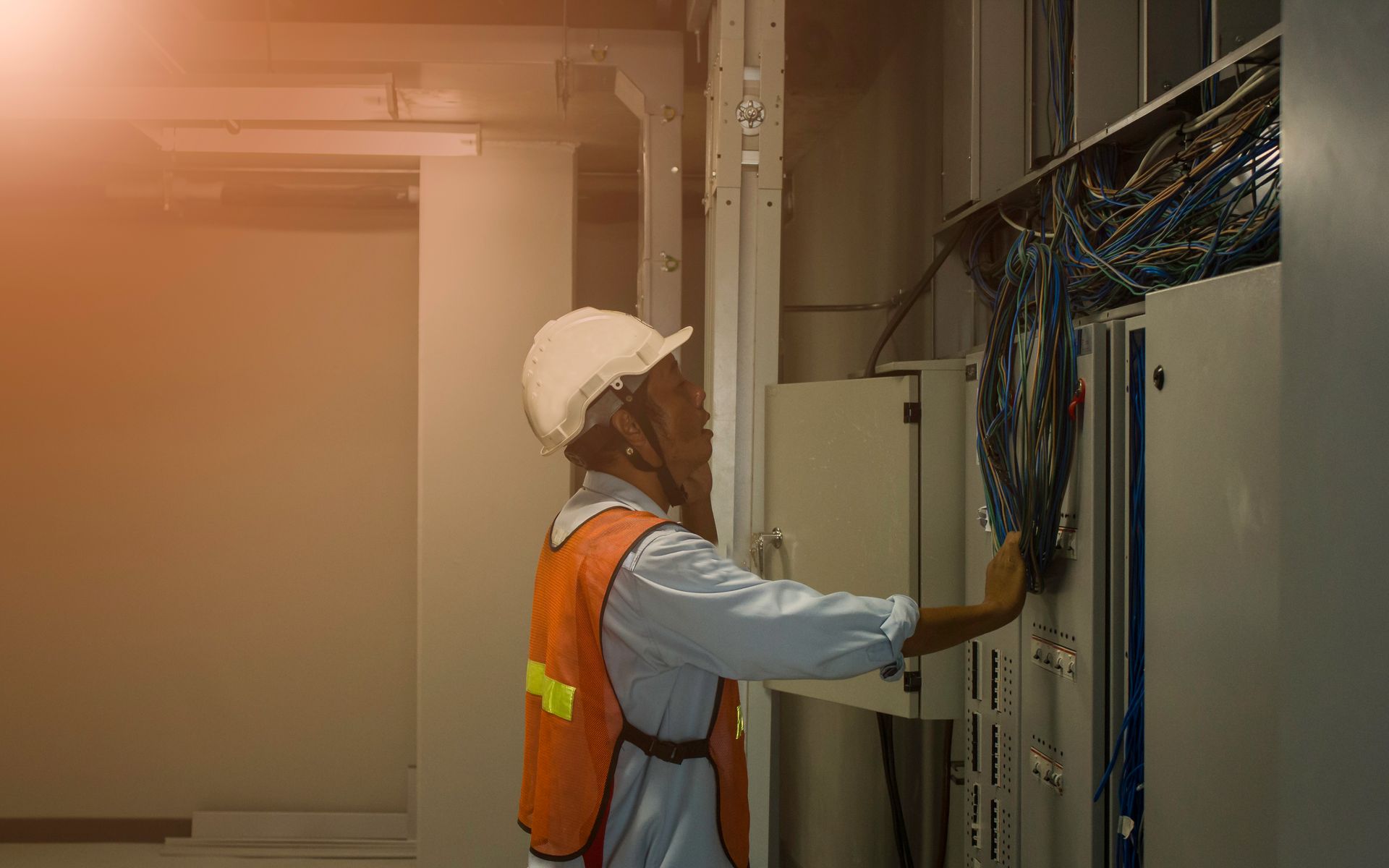 Obrero de la construcción con casco y chaleco inspeccionando cables en un panel eléctrico.