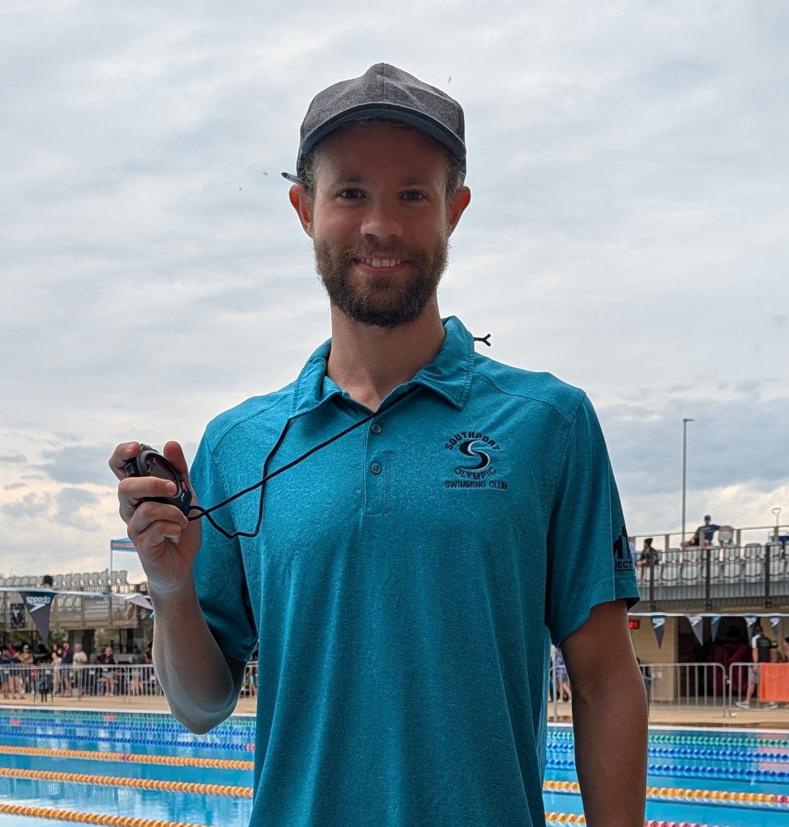 Man in teal shirt, holding stopwatch near a swimming pool.