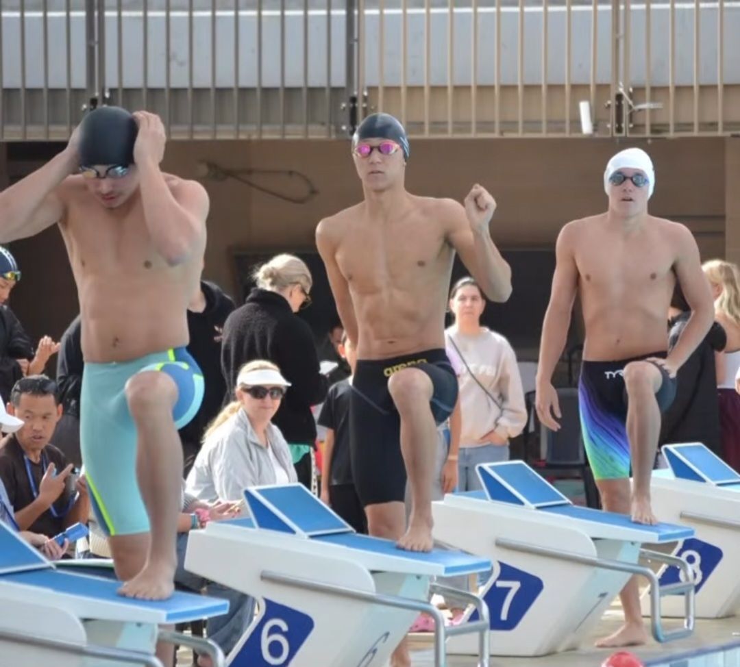 Swimmers in goggles, on blocks, raising arms before a race at a pool.