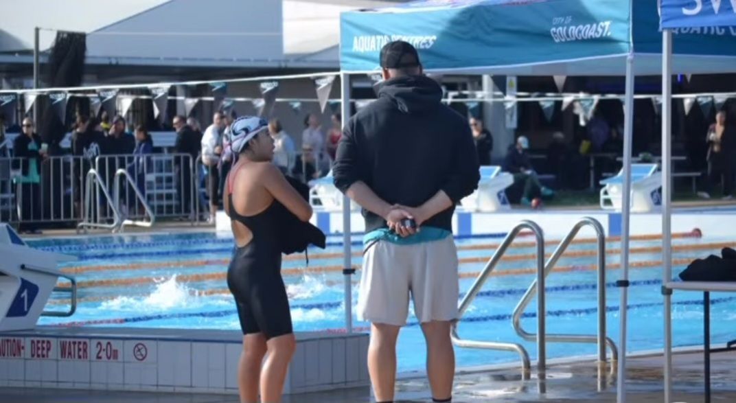 Swimmer in black suit and cap with coach at poolside, watching a race.