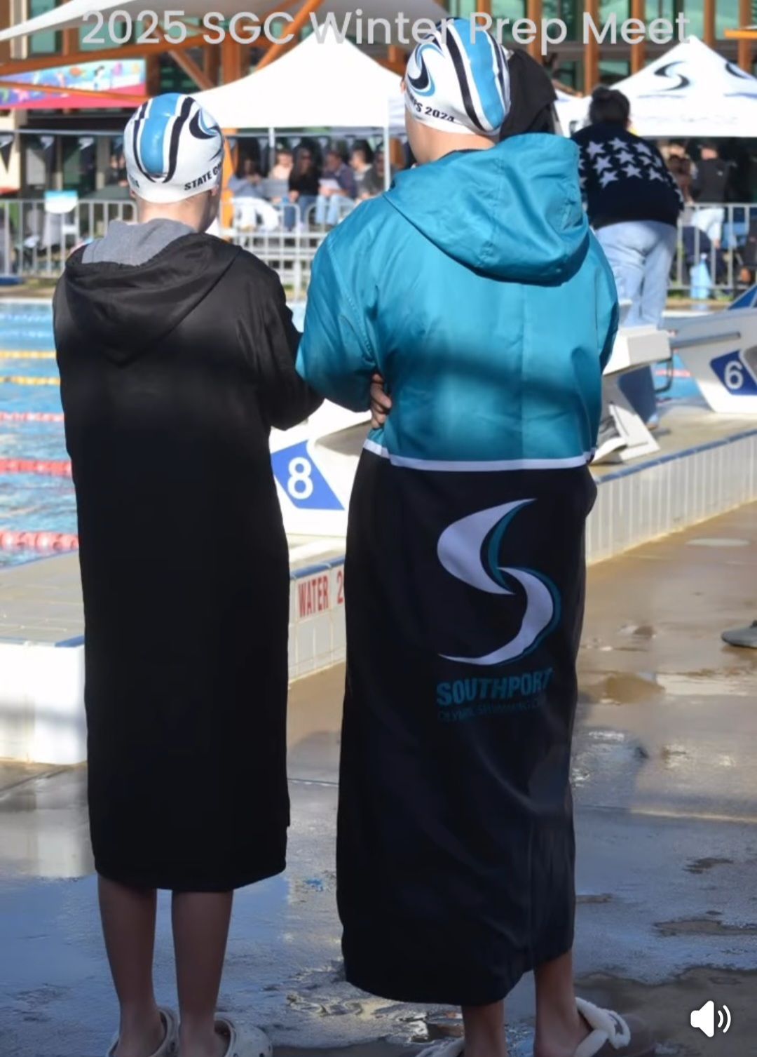 Two swimmers at pool wearing swim caps, hooded robes, and towels, one teal, one black.