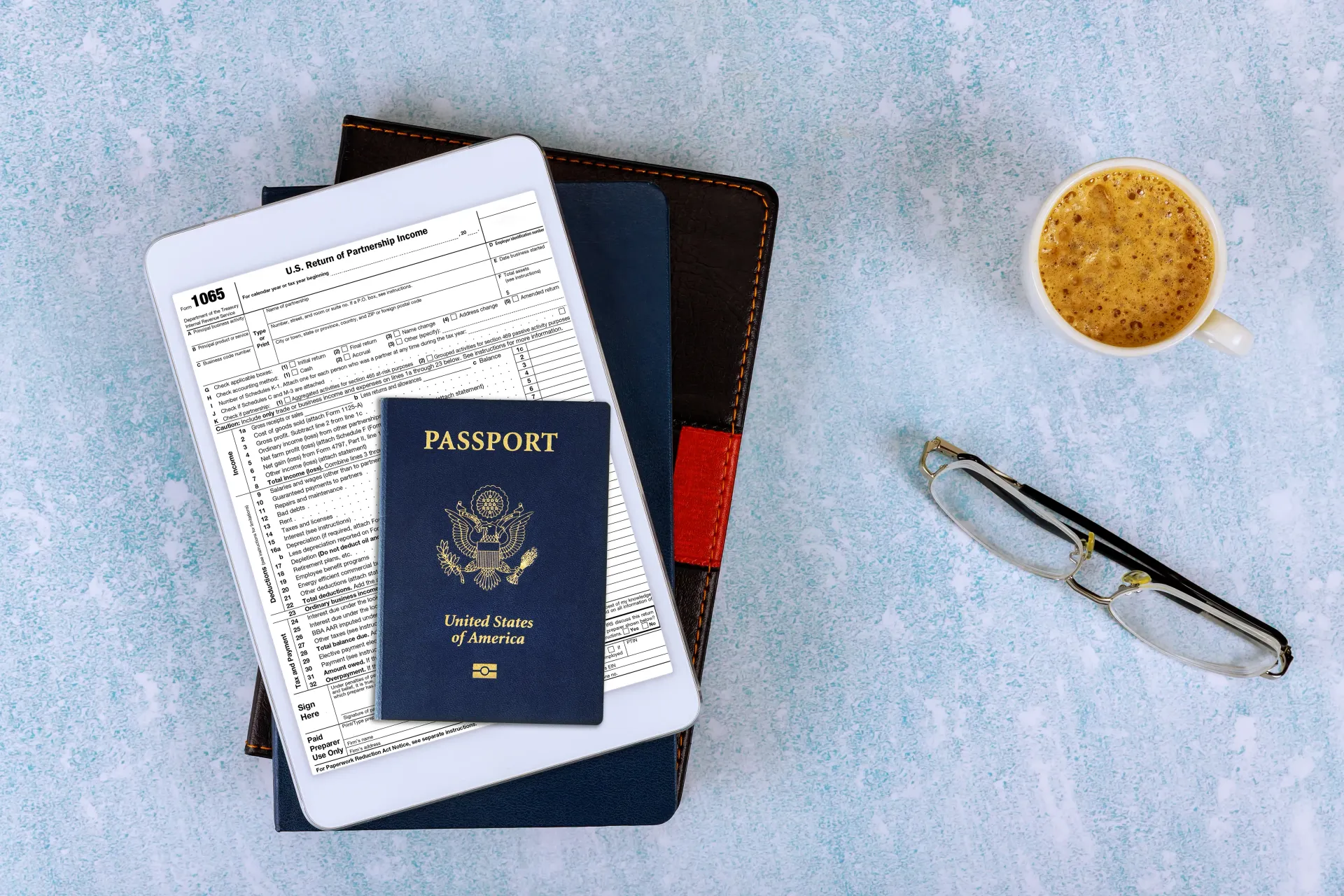 Tablet, passport, books, glasses, and coffee cup on a blue surface.
