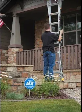 Man on ladder, adjusting it next to a house with stone, brown railing, and ADT sign.