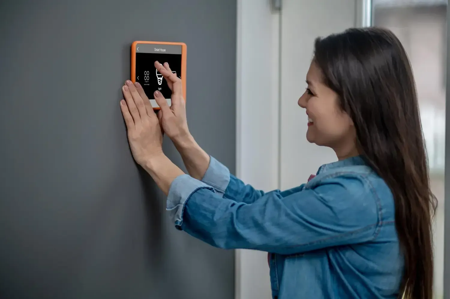Woman adjusting a smart home control panel on a gray wall, smiling in a bright room.