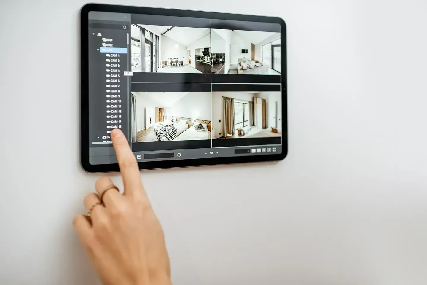 A hand touching a tablet mounted on a white wall, showing four camera views of a home's interior.