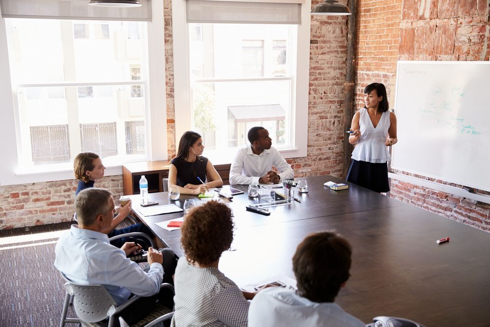 Woman presenting at meeting, writing on whiteboard.  Group seated around table in an office, looking on.