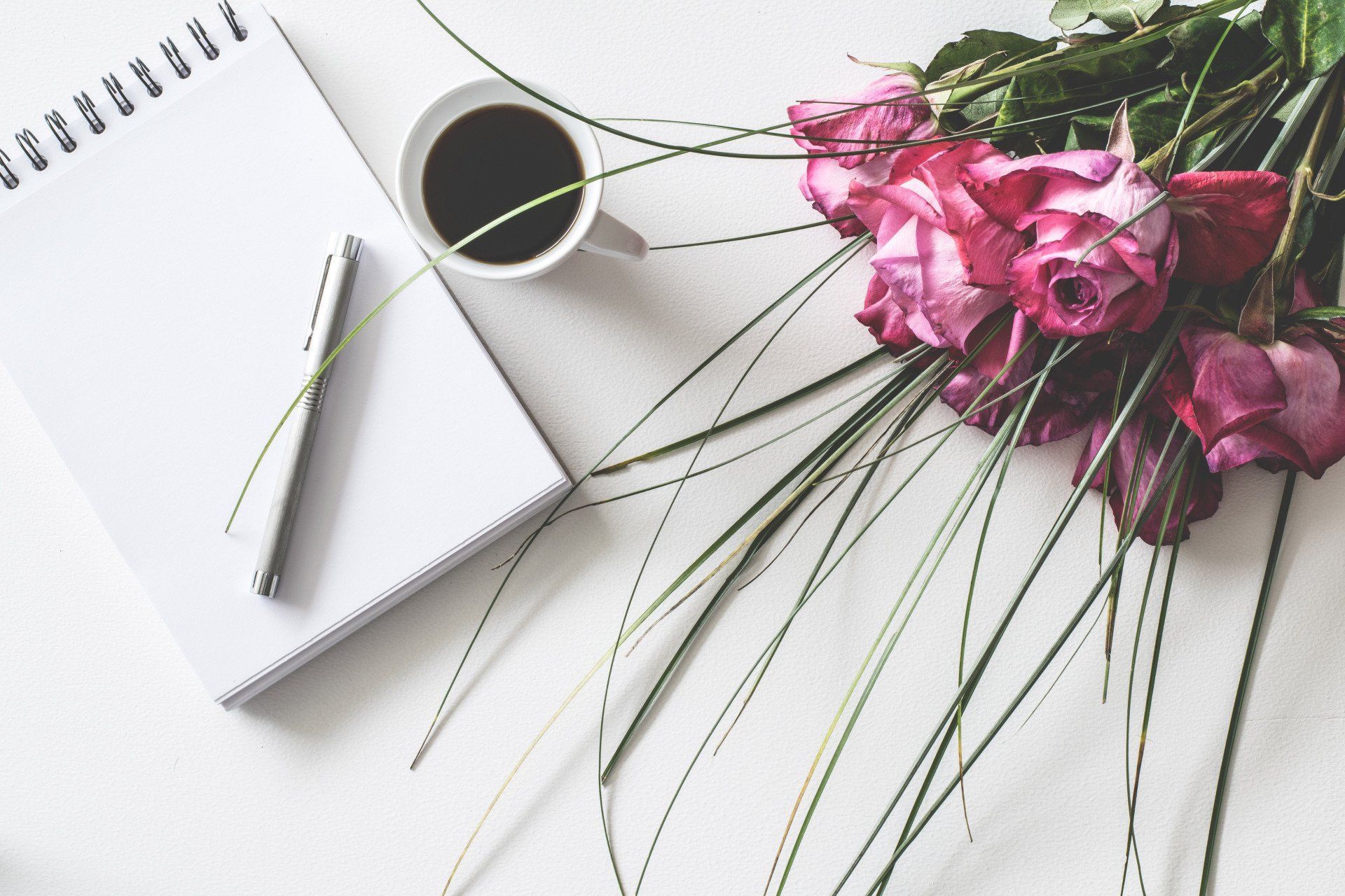Notebook, pen, coffee cup, and pink roses on a white desk flat lay