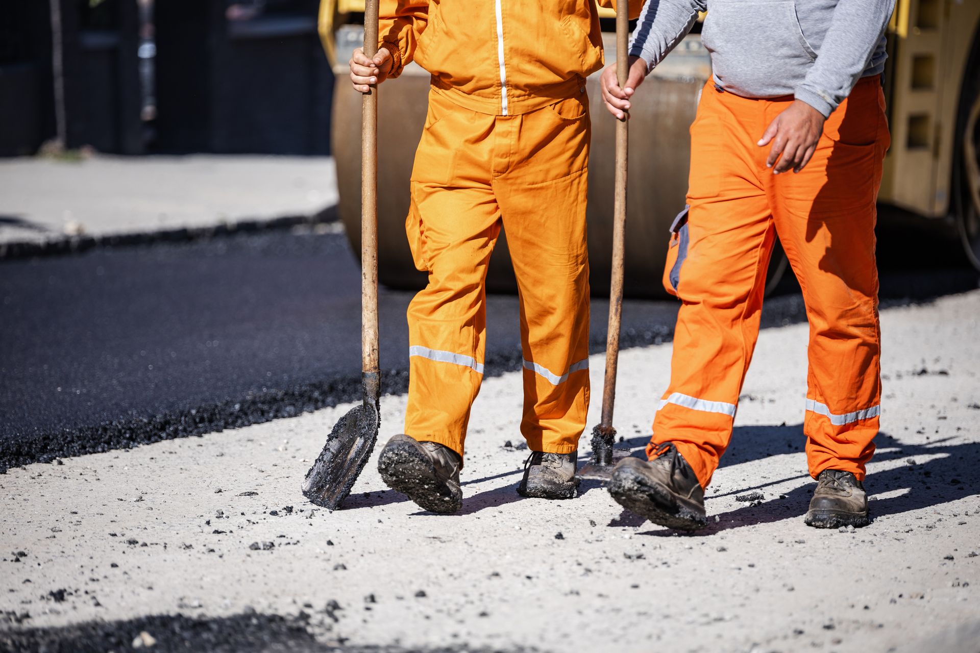 Two workers in bright orange overalls hold tools while standing on a prepared section of a road.