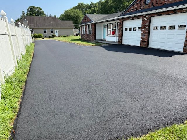 Newly paved asphalt driveway next to a white fence, leading to a brick house with garage doors.