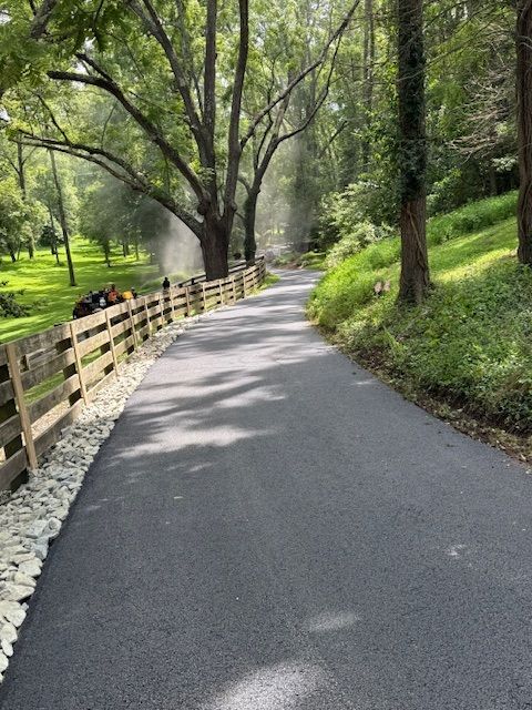 Paved path winding through a wooded area, bordered by a wooden fence and greenery.