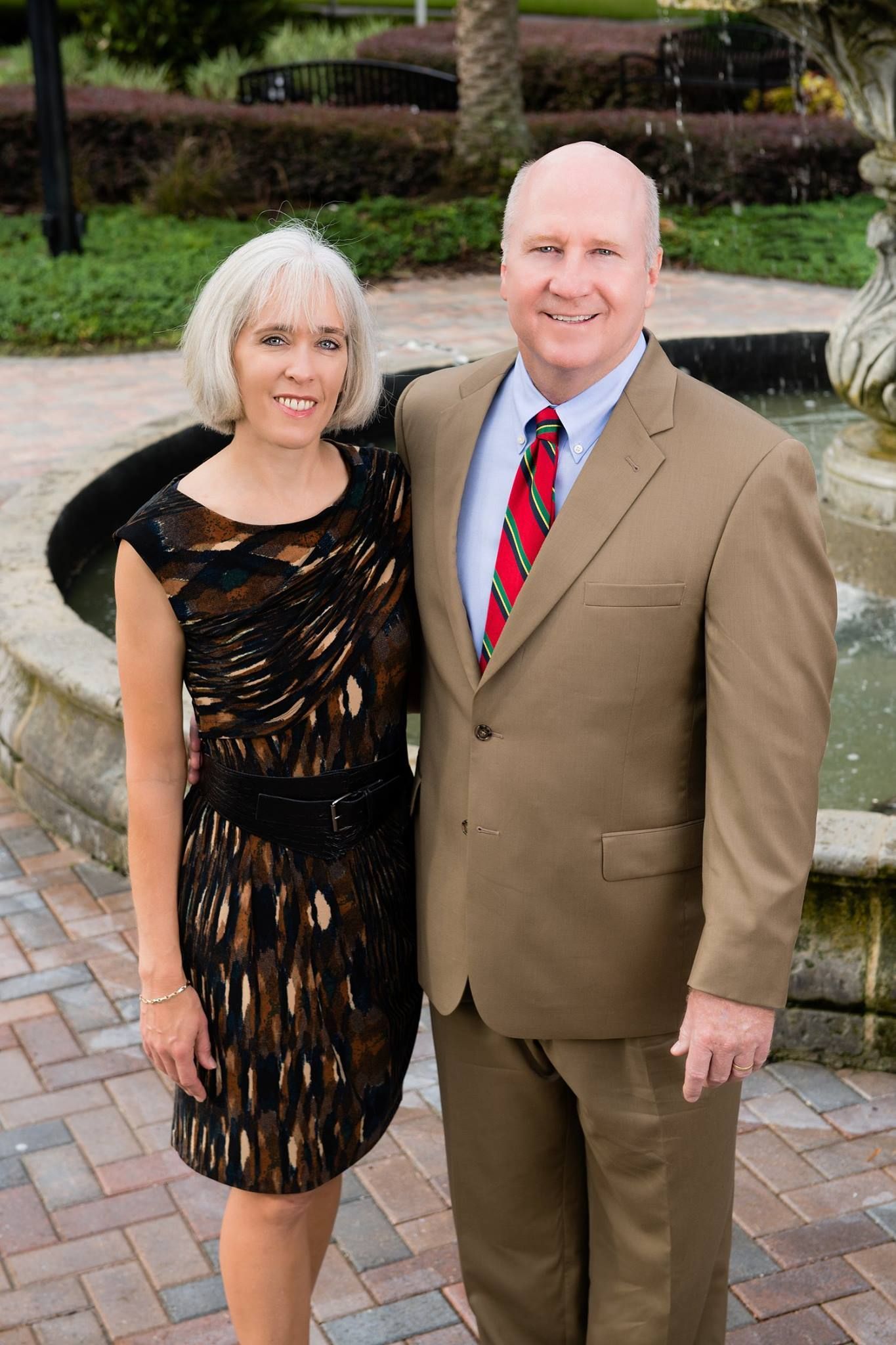 Man in tan suit with woman in patterned dress, smiling, near a fountain.