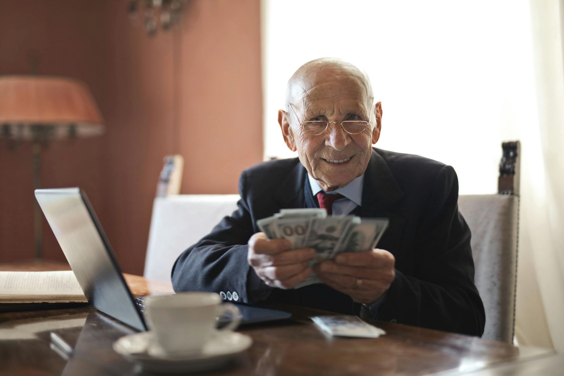 Man in suit counting cash at a table, smiling. Laptop and coffee cup present.