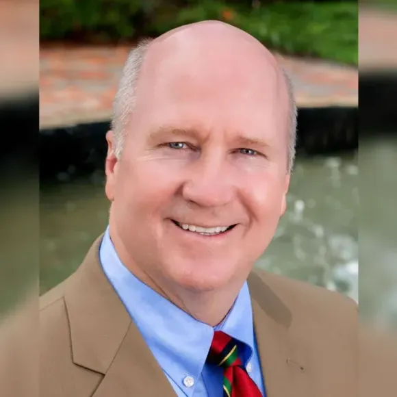 Man smiling, wearing a blue shirt, tan suit jacket, and red tie. Outdoor background.