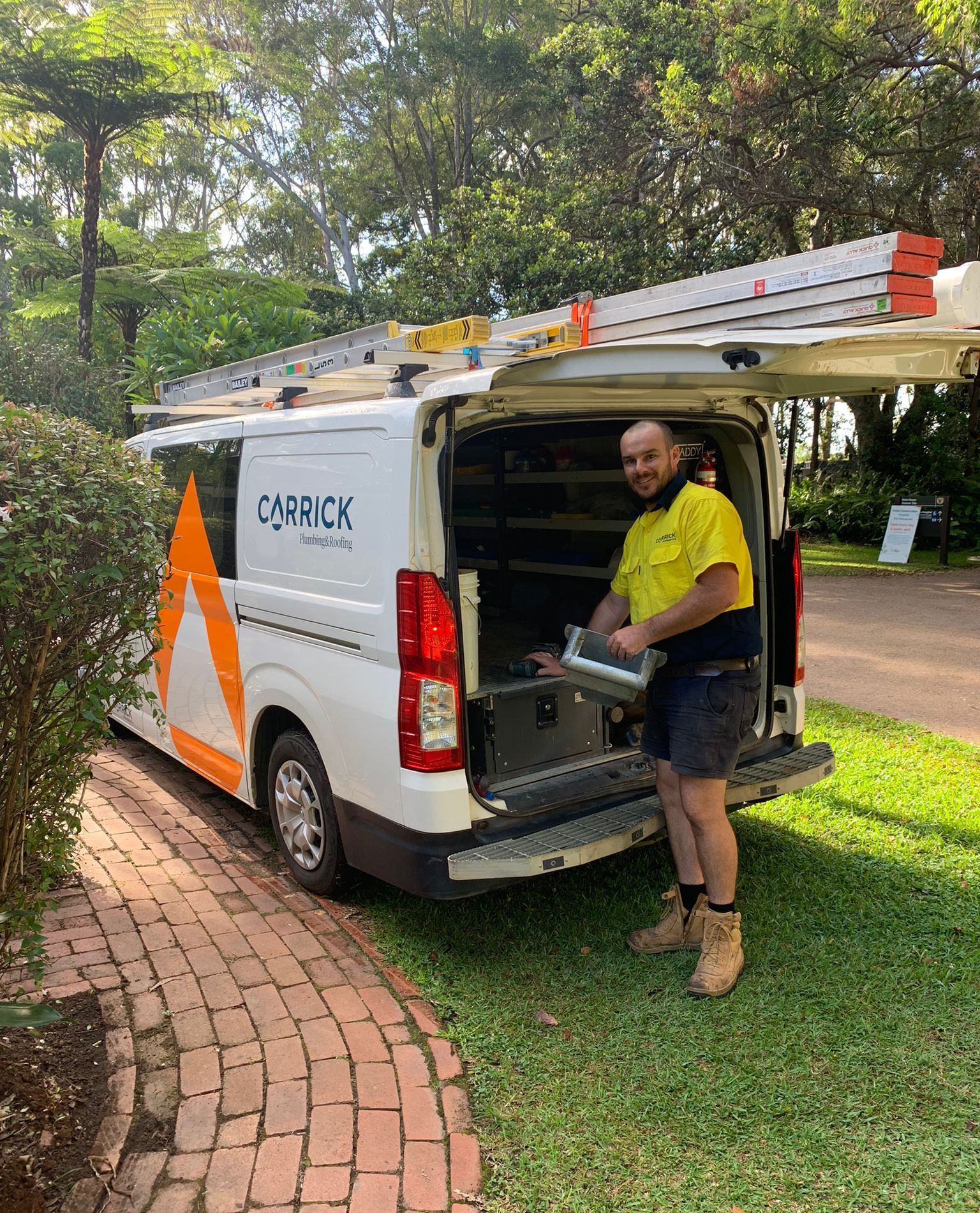 A plumber in work clothes smiles while standing at the back of a van with a ladder on top. The van has a logo