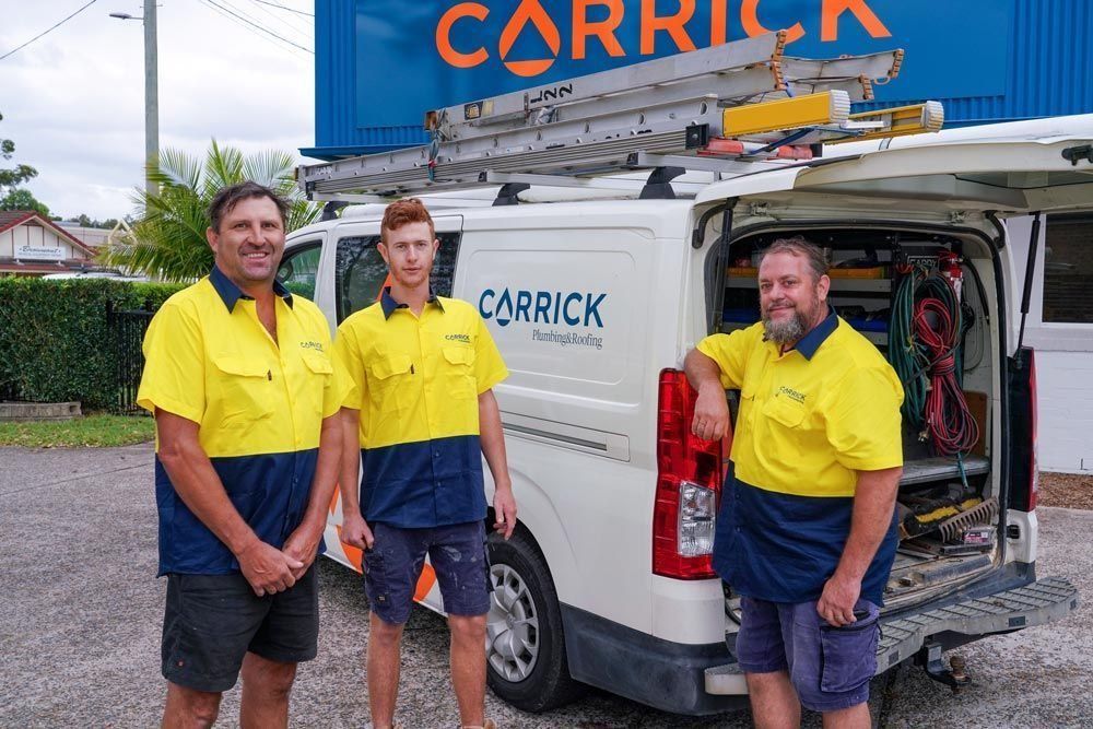 Three men in yellow and blue work shirts stand near a white van with tools. They are in front of a work ute that has the name