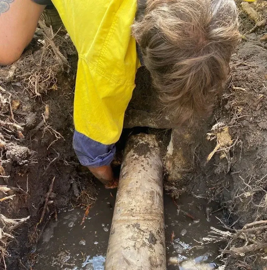 A plumber in a yellow shirt is digging in the dirt near a burst pipe, trying to repair it.
