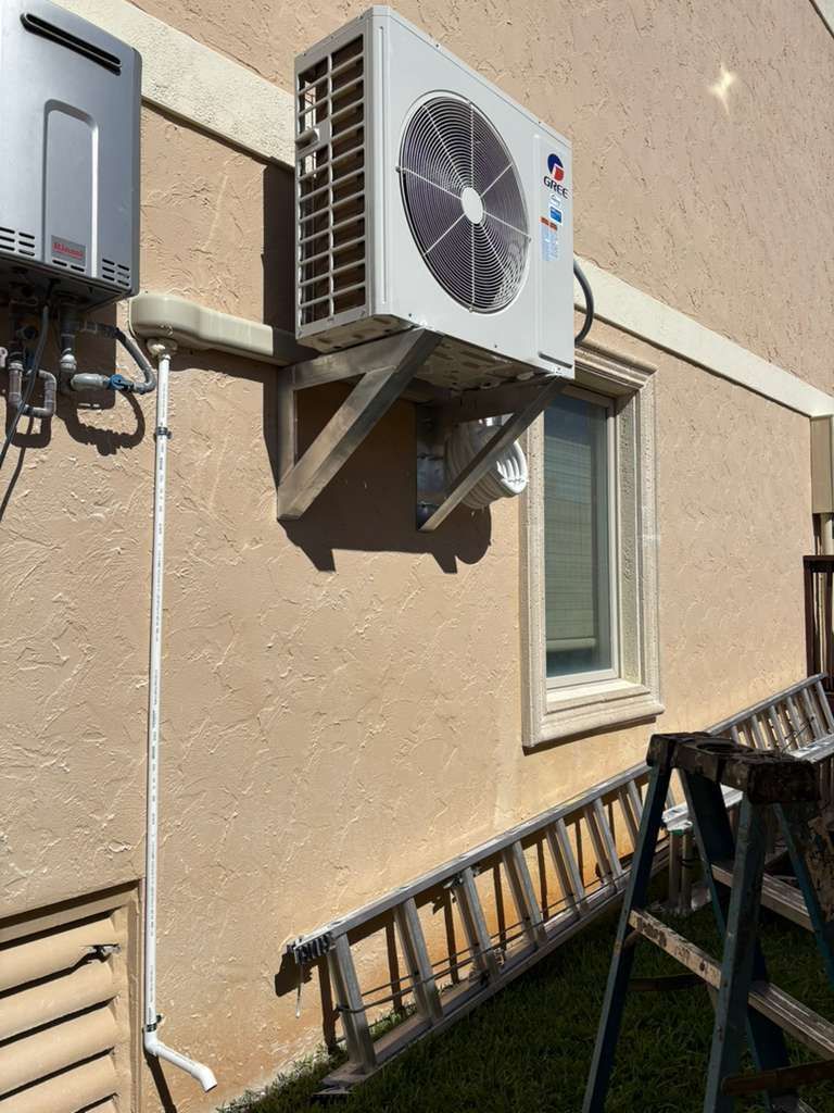 Outdoor air conditioner unit mounted on a white wall beside a window in sunlight