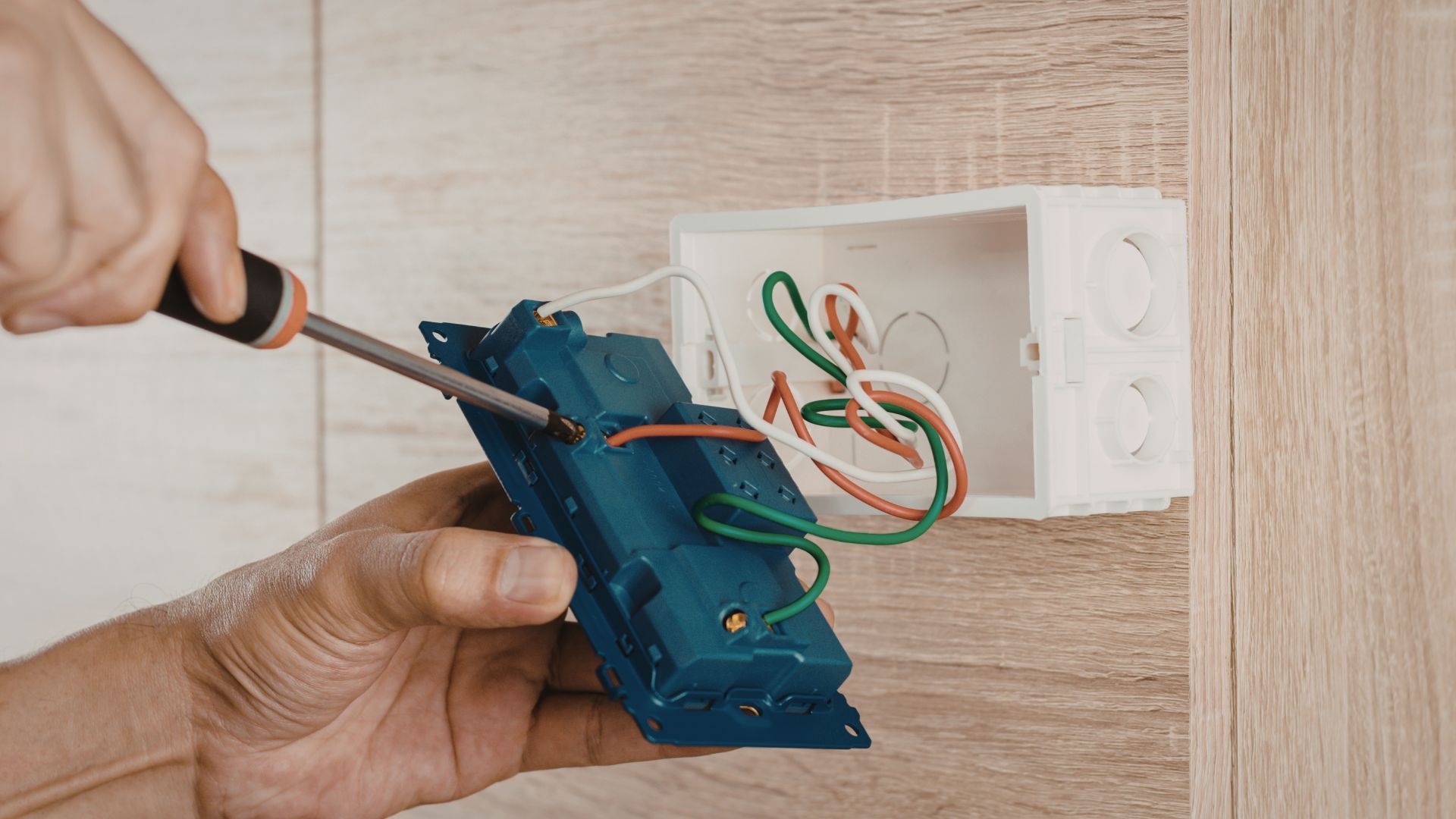 Hands installing a blue electrical outlet box with exposed wires in a wall opening