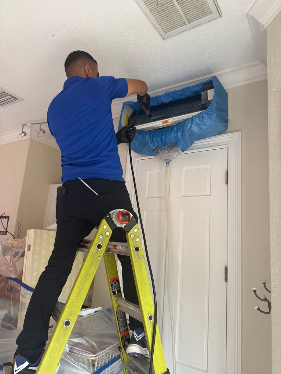 Person on a ladder repairing a blue-covered wall vent near the ceiling in a room