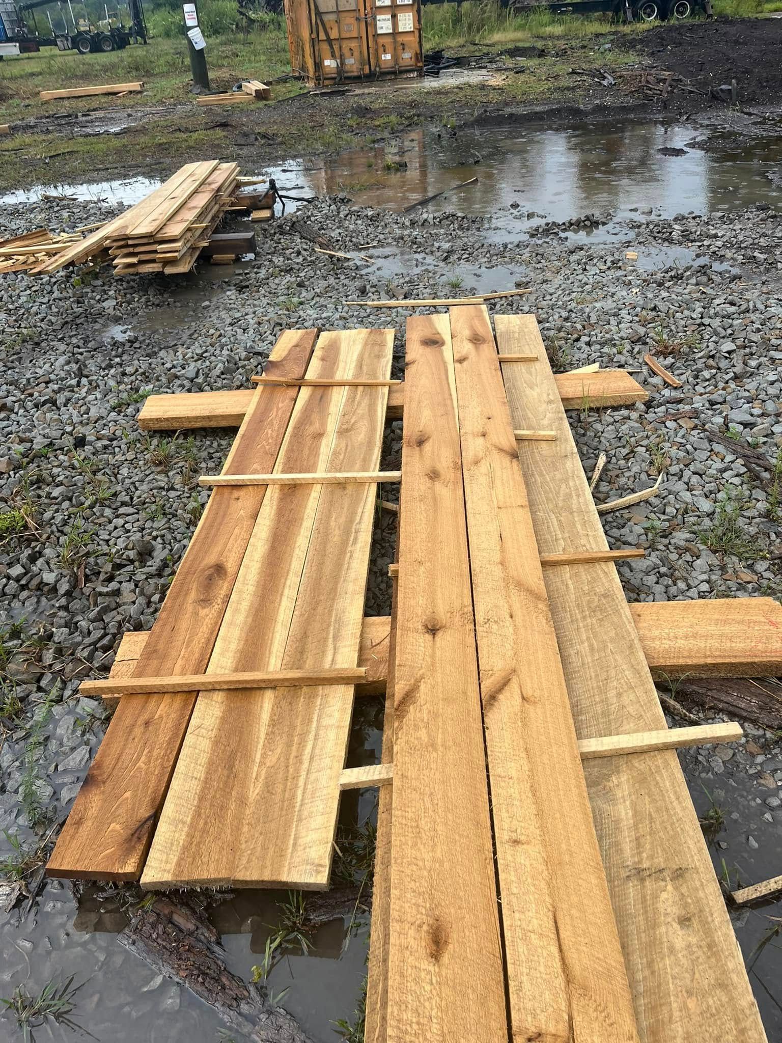 Wooden planks laid across a muddy, wet ground, with more planks and a stack in the background.