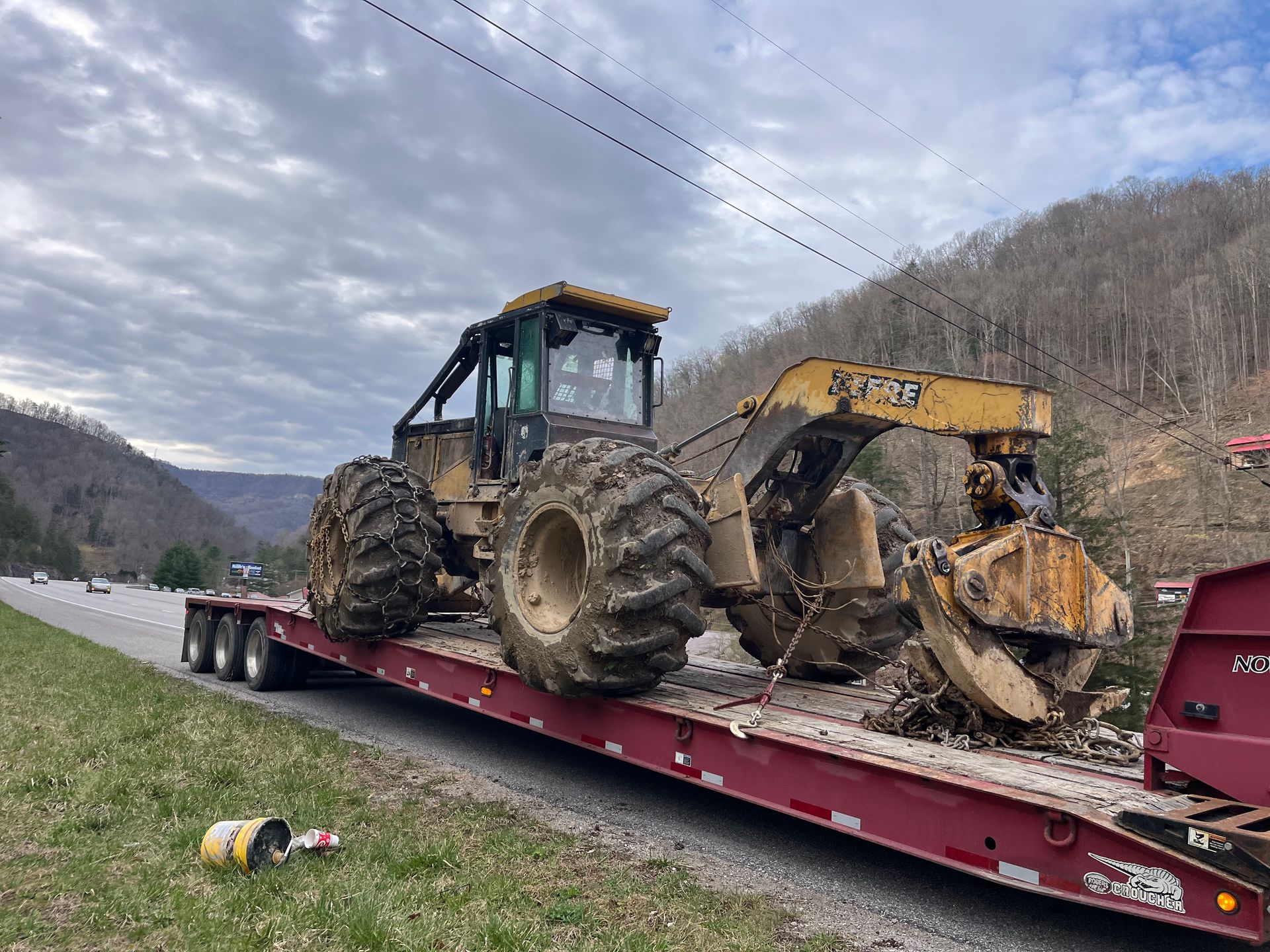 A large, muddy forestry machine on a flatbed trailer beside a highway in a mountain landscape.