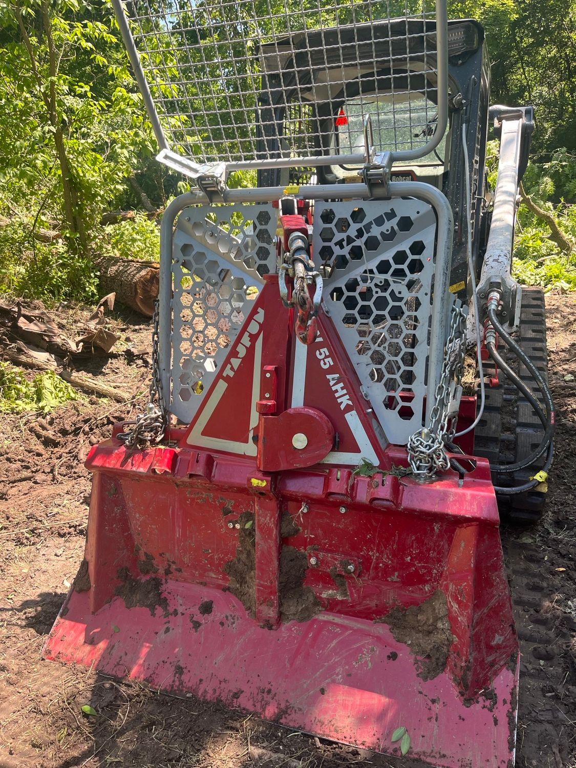 Red and gray forestry mulcher attached to a skid steer in a wooded area.