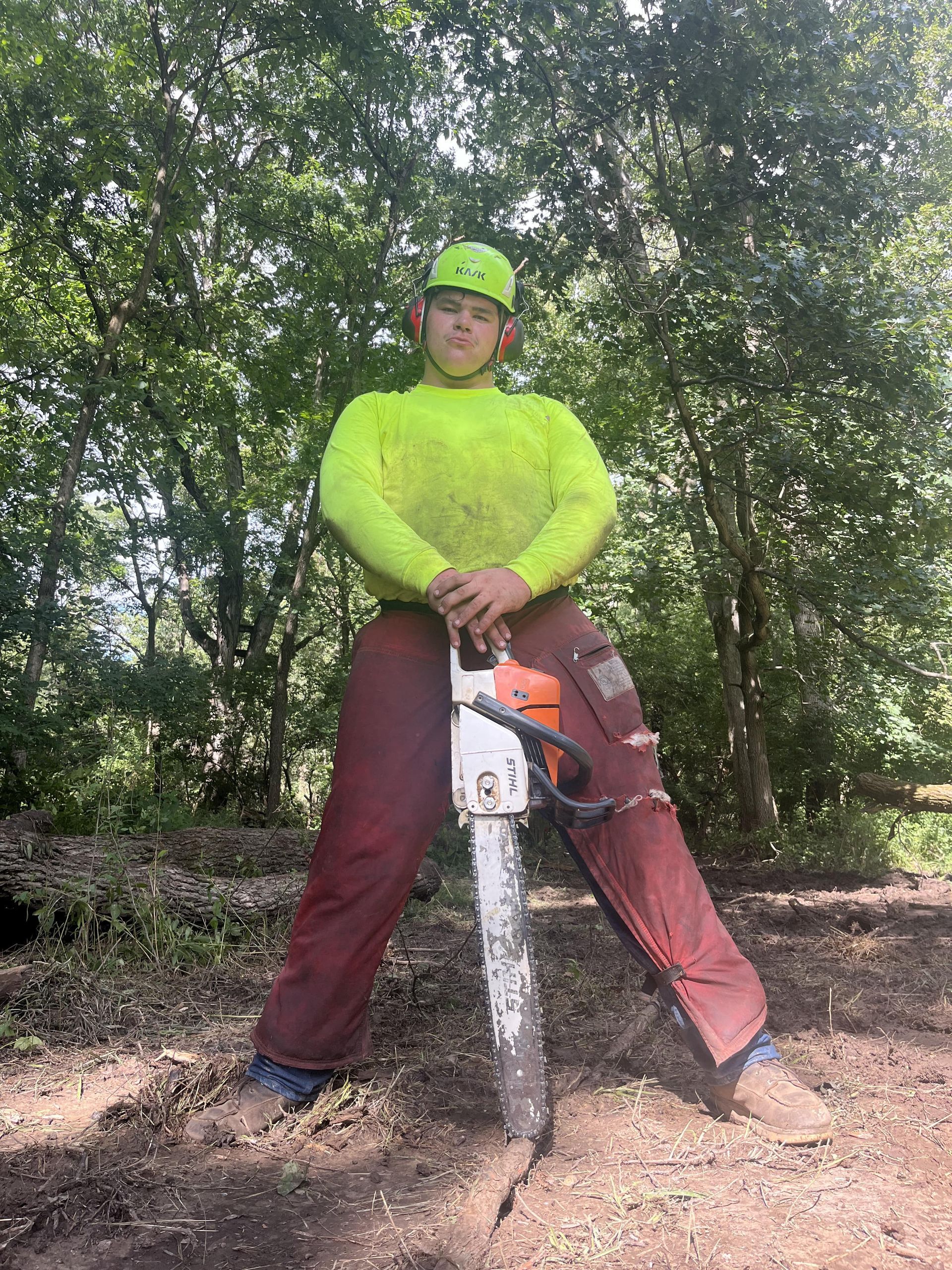 Man in safety gear, holding a chainsaw, standing in a wooded area.