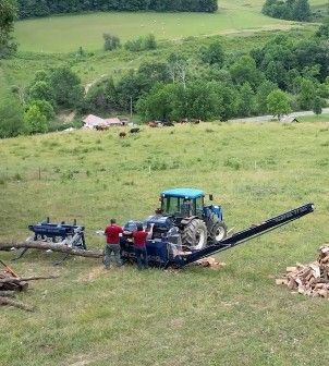 Men using a log splitter attached to a blue tractor in a grassy field with a rural landscape in the background.