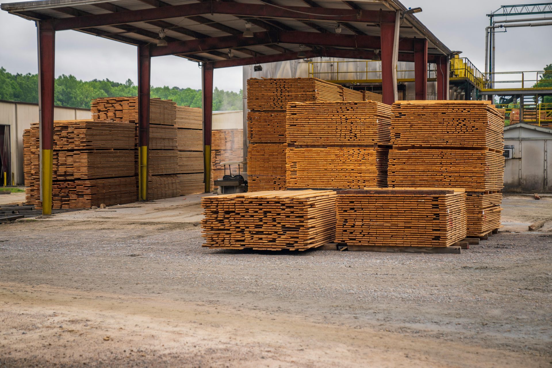Stacks of lumber under a covered shed in a lumberyard, with a cloudy sky in the background.