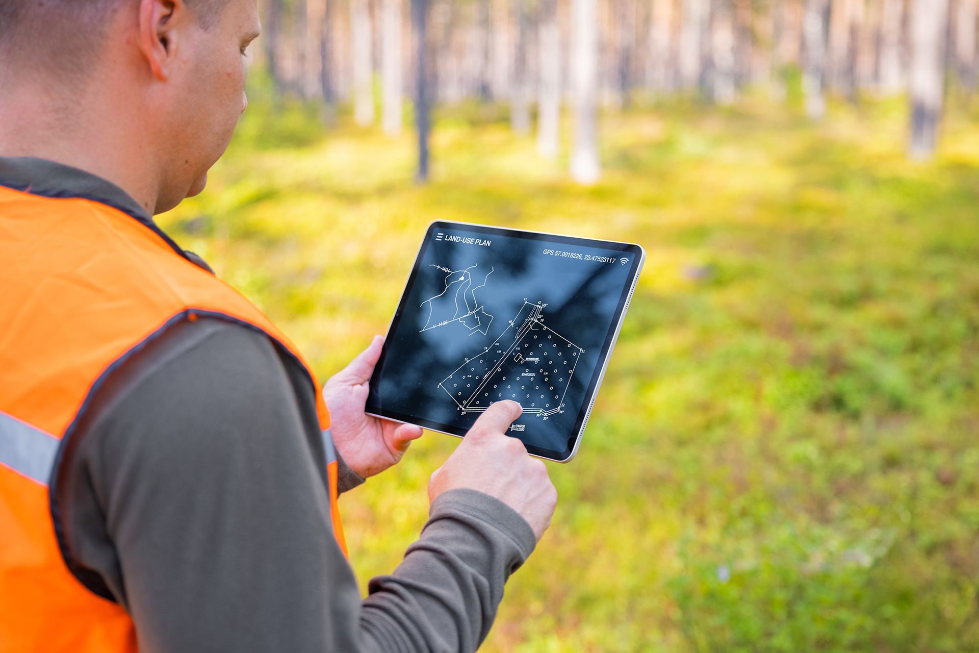 Person in orange vest uses tablet in a forest, displaying a map or data.