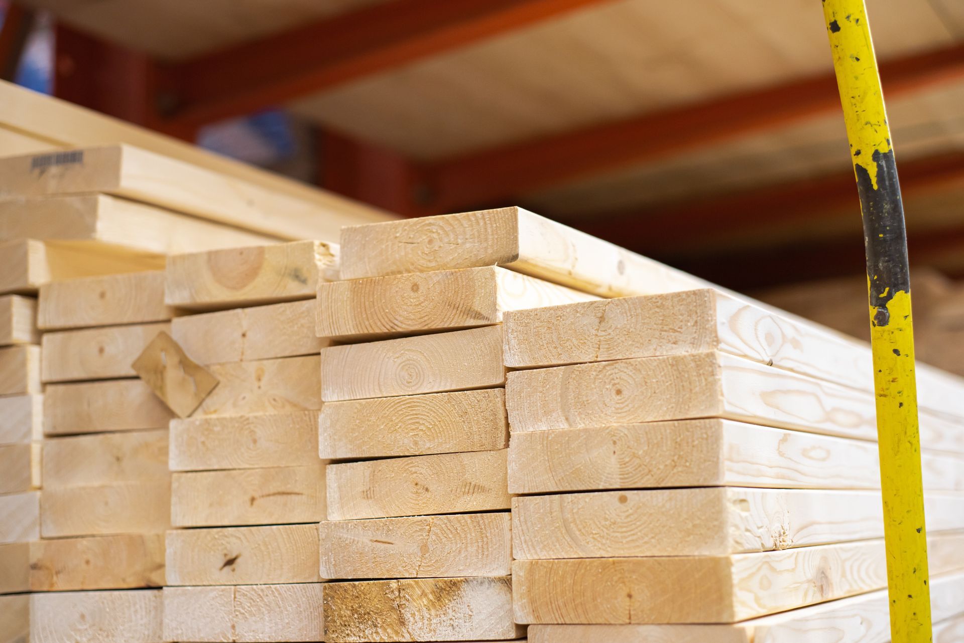 Stack of fresh-cut wooden planks in a lumberyard, with a yellow pole in the foreground.