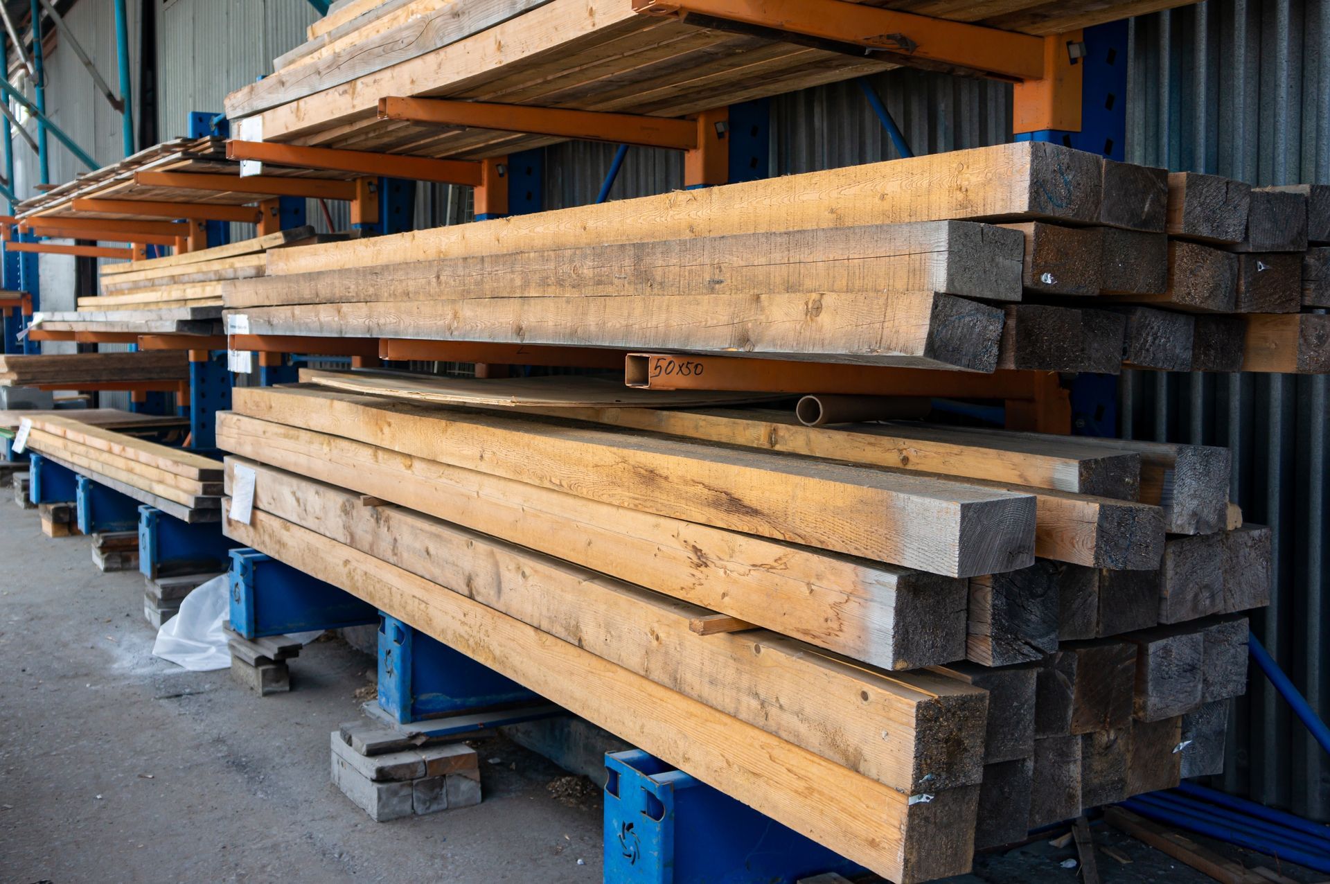 Wooden beams stacked on industrial shelving inside a warehouse. The beams are weathered and rectangular, on blue and orange metal racks.