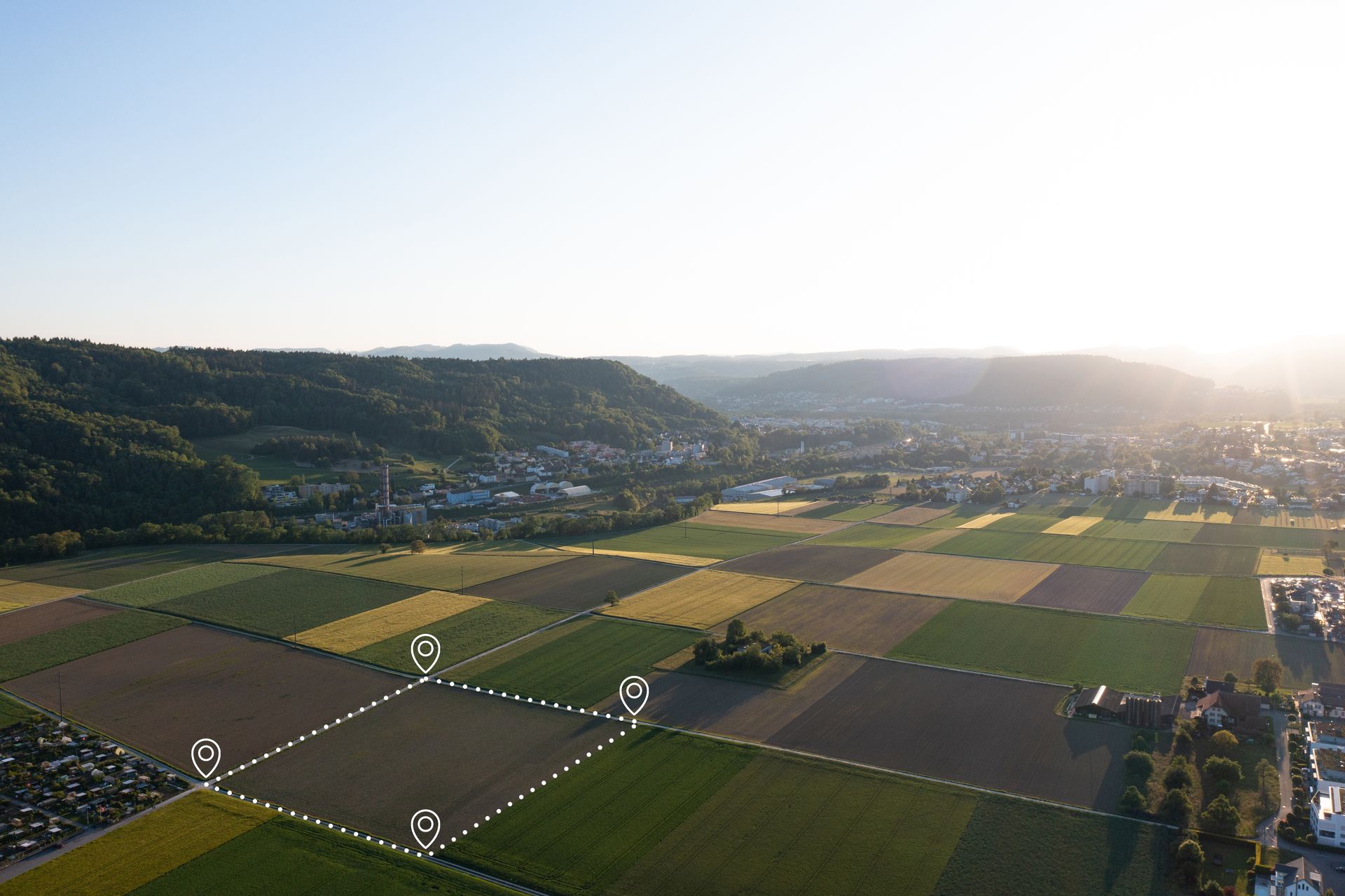 Aerial view of farmland with grid patterns, a town, and mountains under a bright sky.
