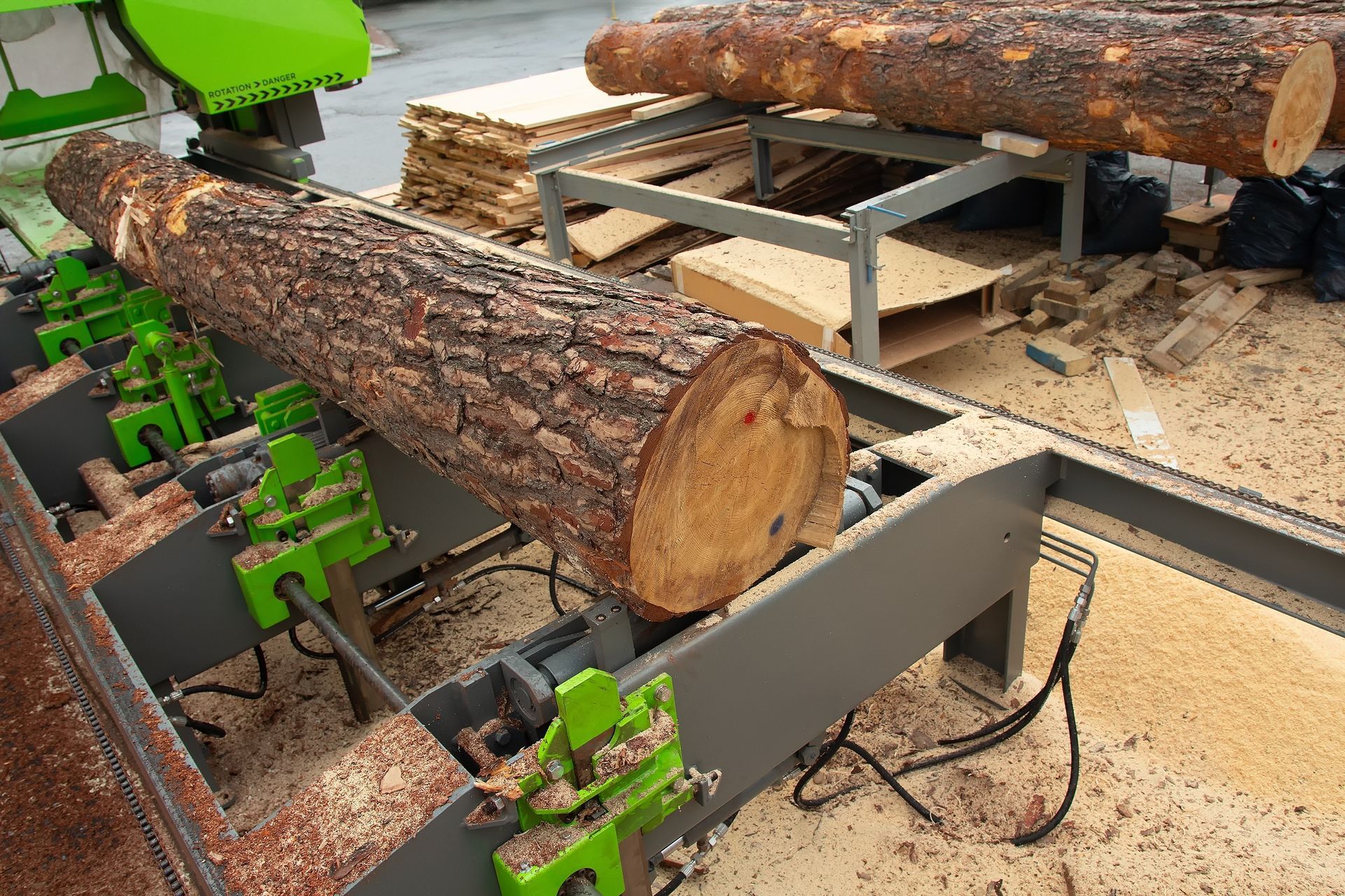 Log being processed on a green and gray industrial sawmill with wood chips and stacked lumber visible.