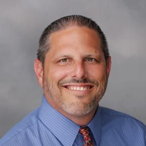 Smiling man with graying hair and beard in a blue shirt and patterned tie.