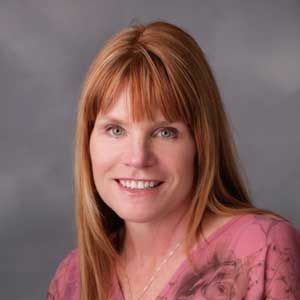 Woman with red hair and bangs, smiling, wearing a pink top, against a gray background.