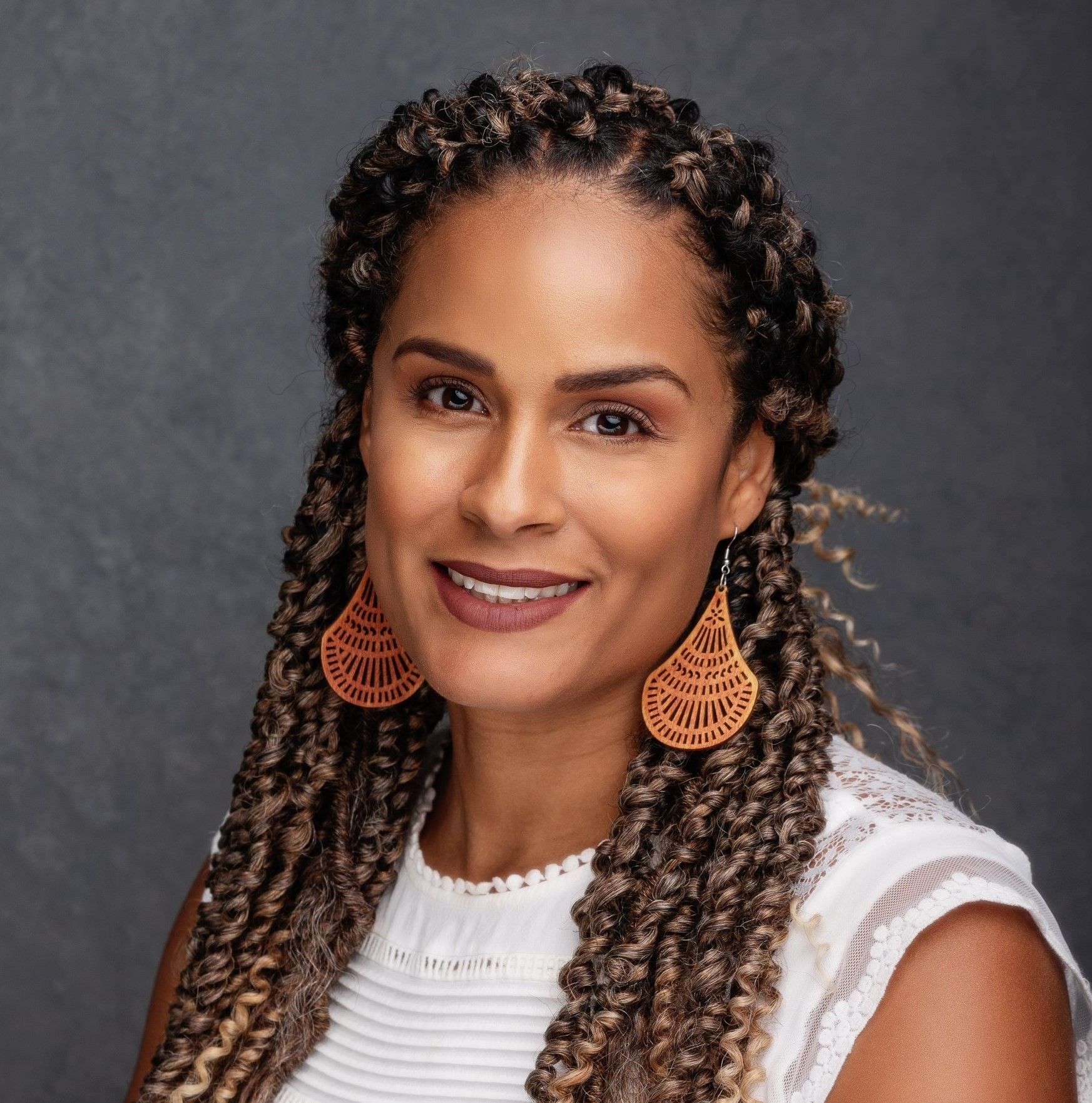 Woman with braided hair smiles, wearing orange earrings and a white top against a gray background.