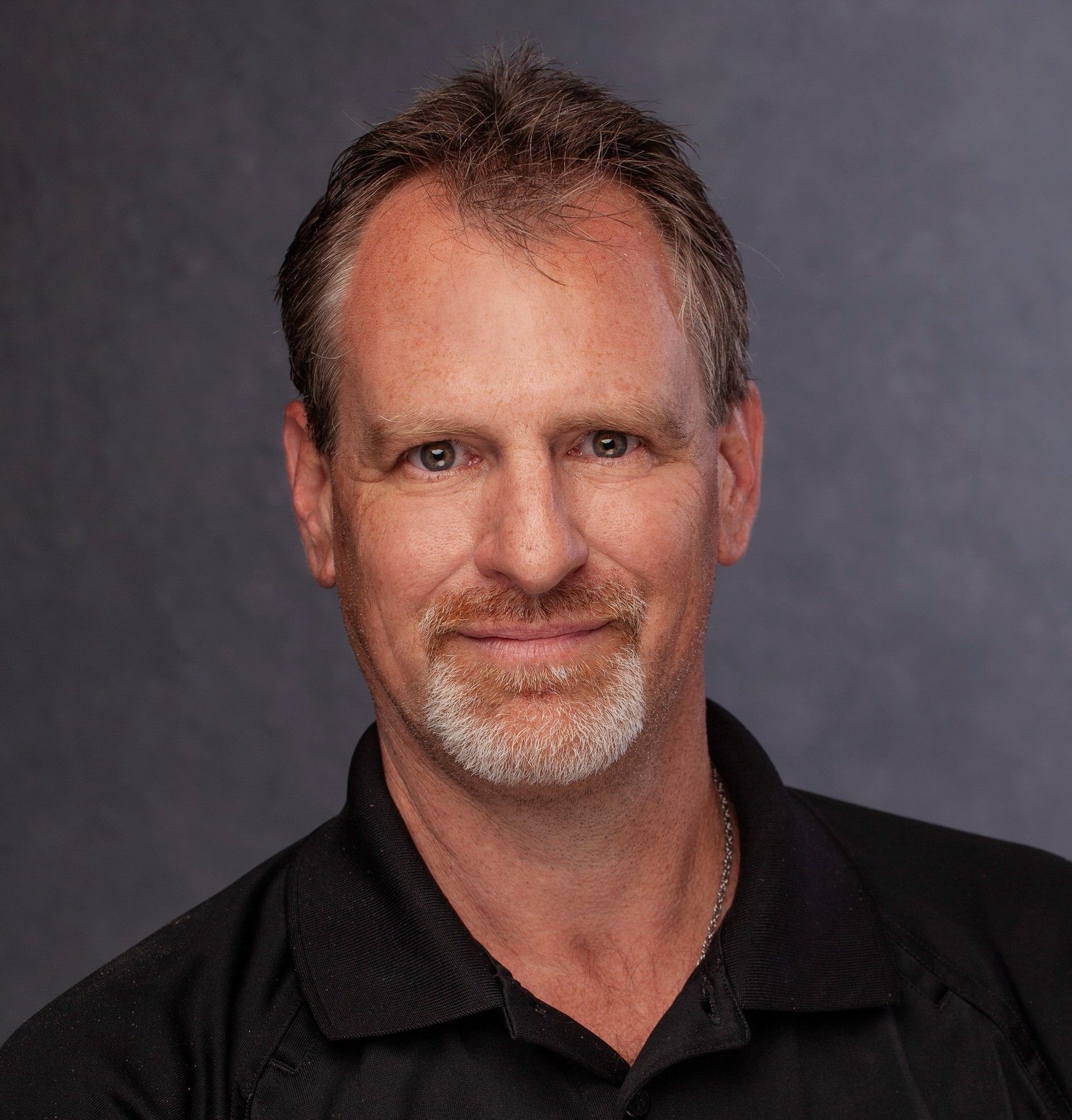 Middle-aged man with graying hair and goatee smiles. He wears a black shirt against a dark background.