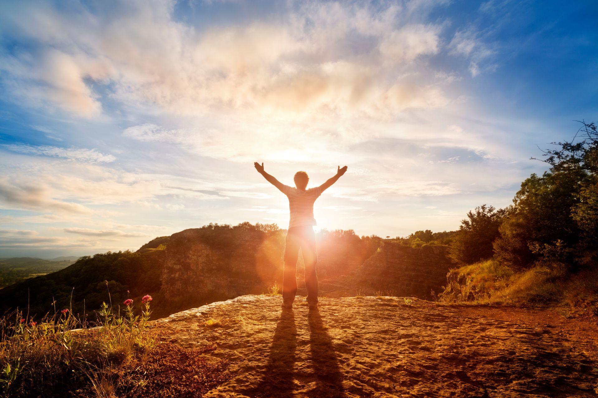 A man is standing on top of a mountain with his arms outstretched at sunset.