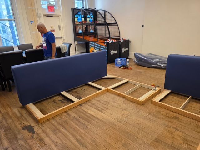 Man assembles blue sectional couch in a room with hardwood floors and a basketball game.