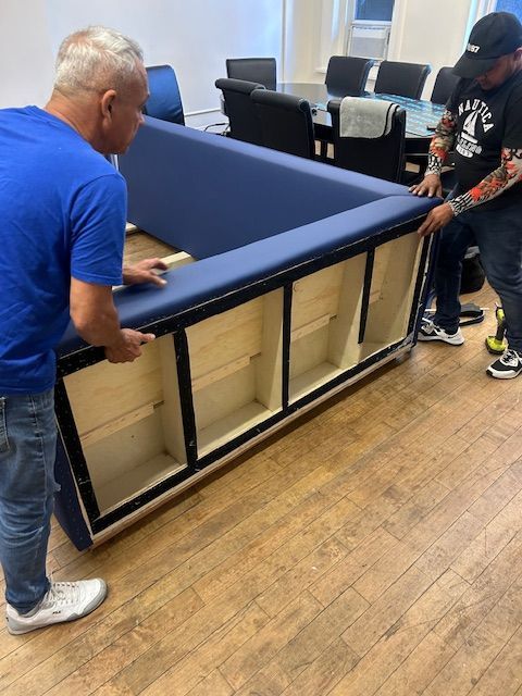 Two men lift a large, blue upholstered furniture piece with wooden shelving in a room.