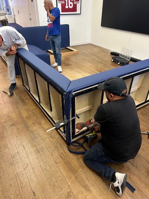 Three men assembling a large, blue sectional sofa on a wood floor. One kneels using tools.