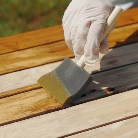 Gloved hand staining wooden planks with a sponge brush; outdoors.