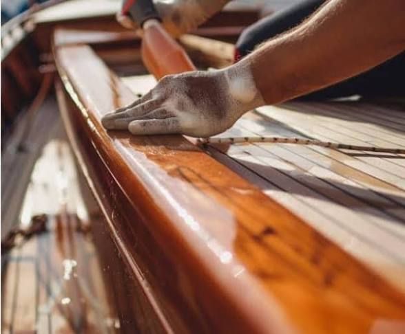 Person sanding the varnished wood railing of a boat, hands in gloves, outdoors.